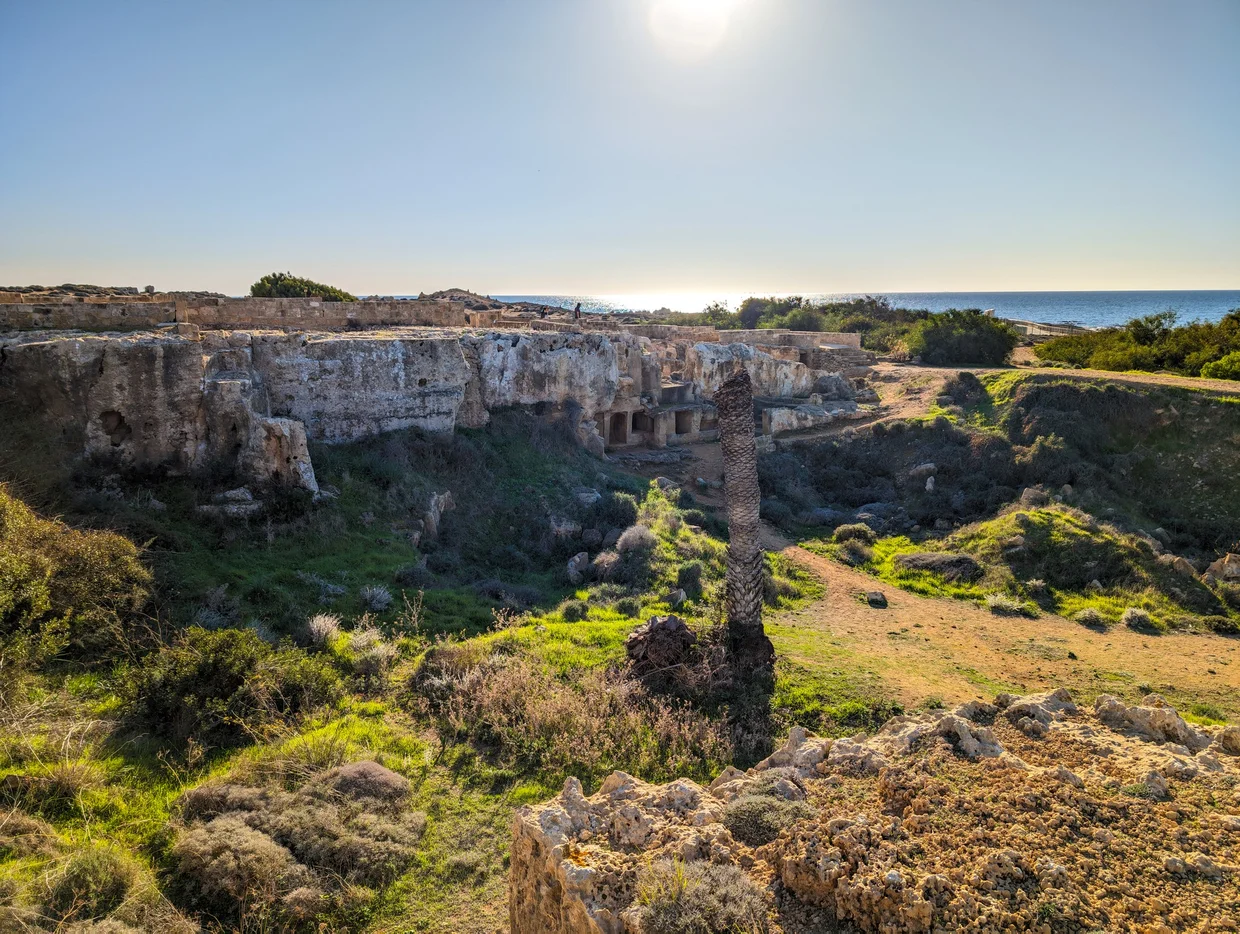 Overview of a sunken tomb complex with overgrown vegetation and sea in the distance