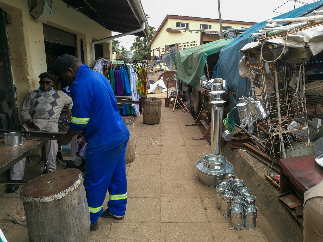 Market alley in Mbabane with a worker, clothing racks, and metalware stalls