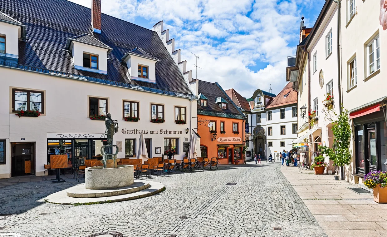A quiet cobbled square in Fuessen's old town with a stone fountain, cafe tables and painted gable houses