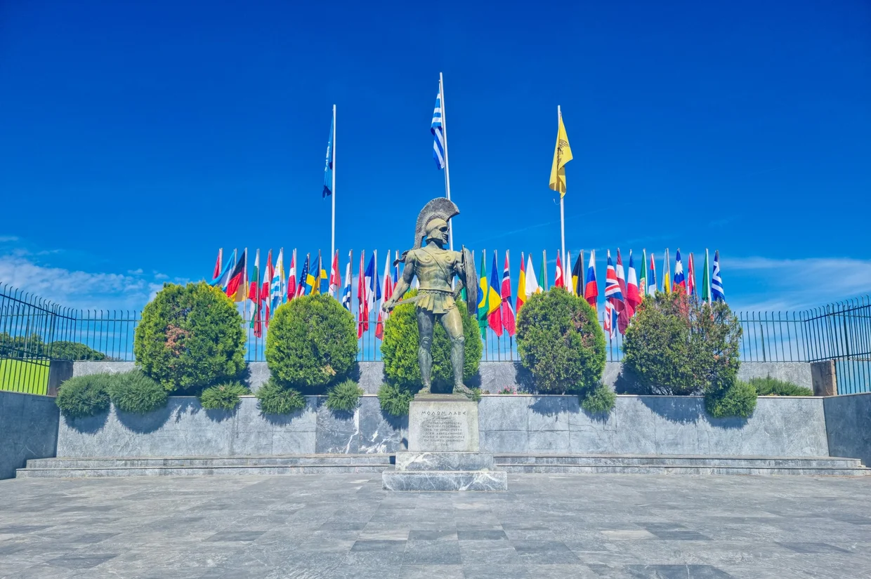 Bronze Leonidas statue in Sparta, holding a shield and spear, with international flags behind
