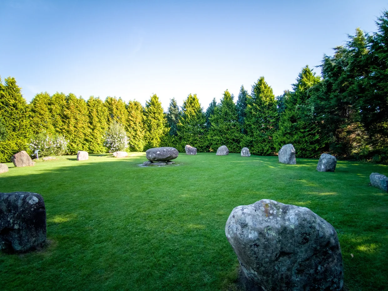 Standing stones arranged in a circle on a green lawn surrounded by trees
