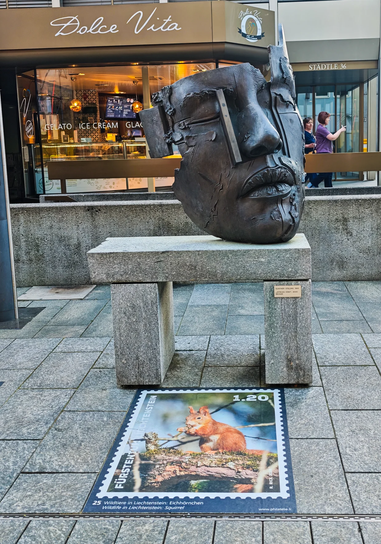 Bronze face-mask sitting on a granite bench, with a wildlife stamp printed on the pavement