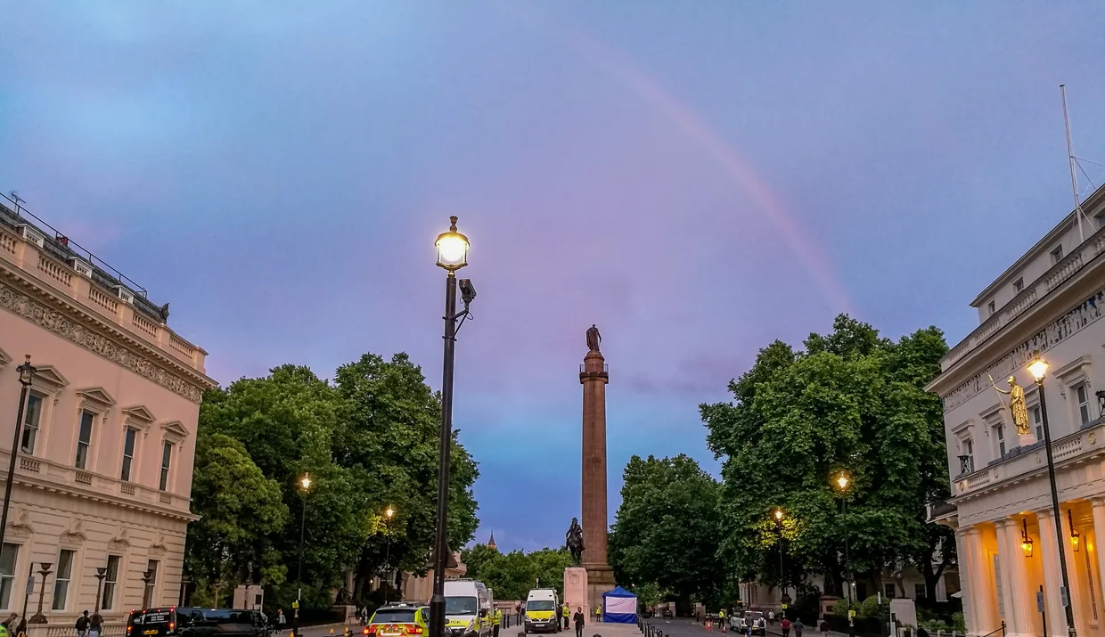 A rainbow arcing over Waterloo Place at dusk, lampposts glowing, a column silhouetted against the pink sky